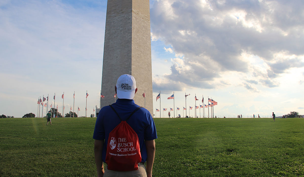 Student wearing Busch School of Business hat and backpack in front of Washington Monument