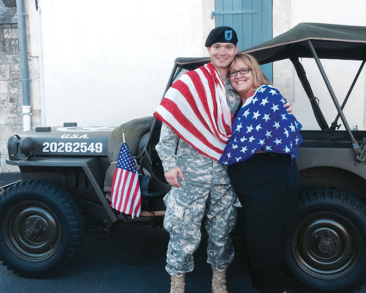 American soldier and his mother draped in an American flag standing in front of a military Jeep