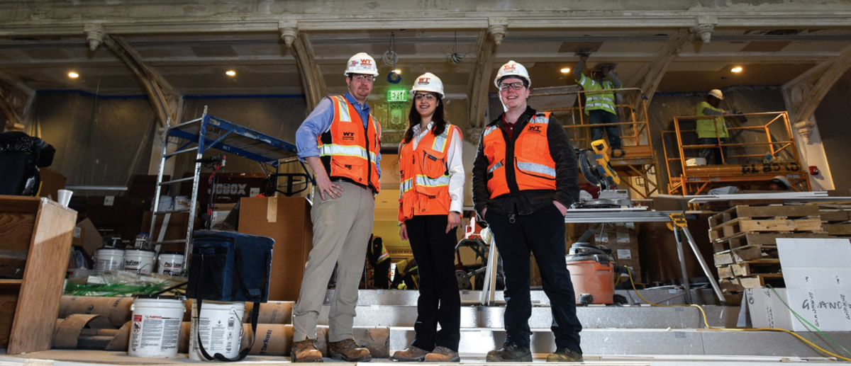Three CUA alumni who worked on the project pose in hardhats in auditorium during the renovation