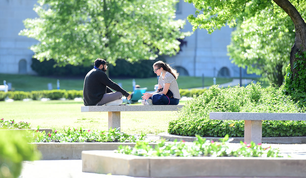 students sit on a bench in a plaza on campus surrounded by trees
