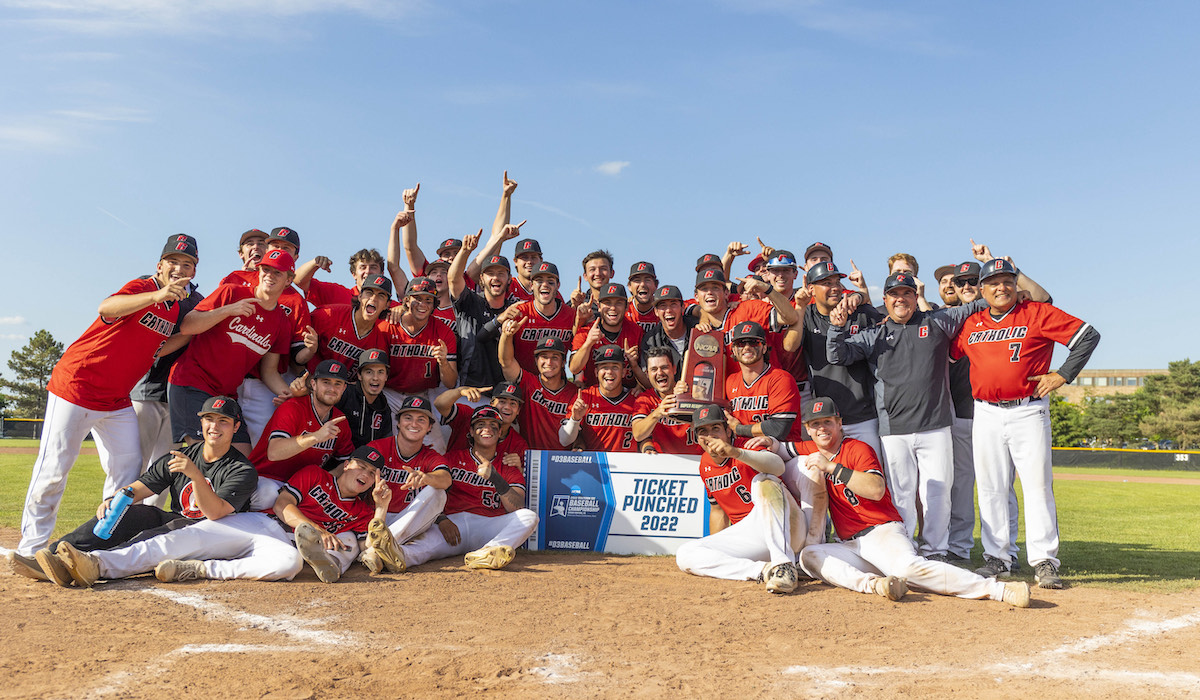 the cua baseball team celebrates their Super Regionals win posing for a picture around a commemorative banner