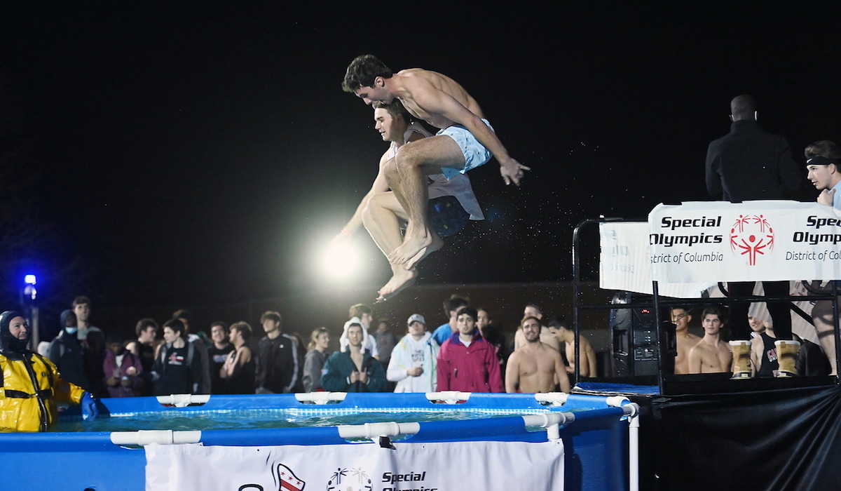 Two male students in swim trunks mid-jump into the pool.
