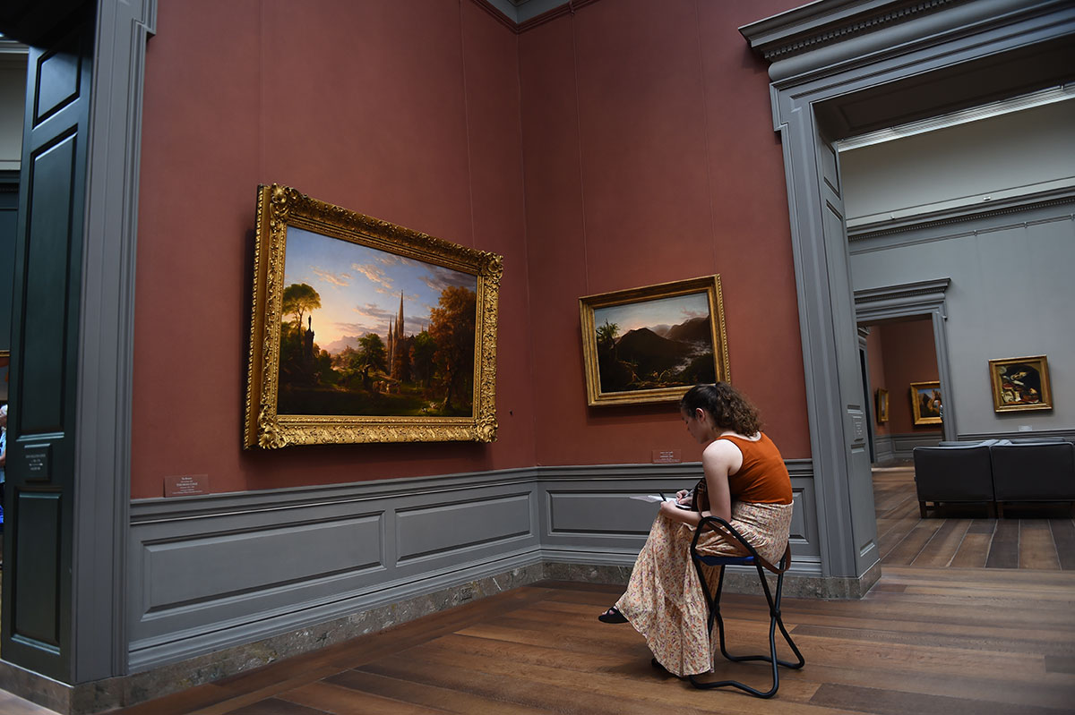 Student sitting on chair in art museum, looking at artwork and drawing